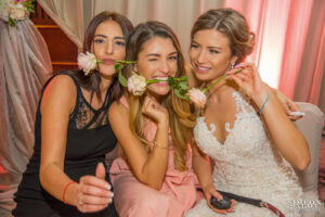Булката Саня и нейните приятелки позират за забавна снимка, държейки рози в уста, по време на голямото сватбено тържество в хотел Маринела, кадър на професионален сватбен фотограф Симеон Сълов.