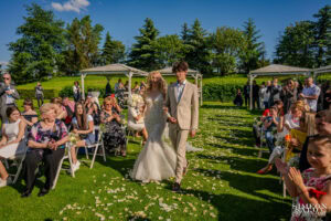 Сияеща булка върви по пътеката, посипана с листенца, към олтара, съпроводена от своя син, докато гостите ръкопляскат на сватба в Голф клуб "Св. София". Кадър от сватбен фотограф Симеон Сълов.