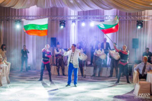 Народният певец Николай Славеев пее на сцена, заобиколен от танцьори, развяващи български знамена, по време на фолклорната програма на сватбата, кадър на професионален сватбен фотограф Симеон Сълов.