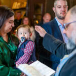 Свещеникът дава своята финална благословия над малкия Борис, който е в ръцете на майка си, в края на ритуала по светото кръщение. Кадър от професионален фотограф за кръщене Симеон Сълов.