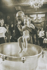 Свещеникът държи малкия Борис над купела по време на ритуала по светото кръщение, улавяйки емоционалната реакция на детето. Кадър от професионален фотограф за кръщене Симеон Сълов.