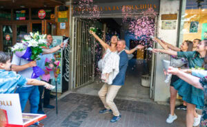 Младоженецът Марио носи на ръце булката Мирела при излизането им от ритуална зала Триадица, докато гостите ги обсипват с конфети. Кадър от професионален сватбен фотограф Симеон Сълов.