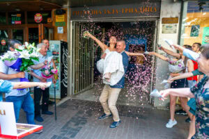 Новото семейство Мирела и Марио излизат от ритуална зала Триадица под дъжд от конфети, носени на ръце от младоженеца. Кадър от професионален сватбен фотограф Симеон Сълов.