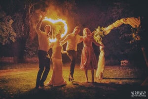 Младоженци и кумове се забавляват и танцуват пред голямо пламтящо сърце, артистичен кадър от сватбено тържество, заснет от сватбен фотограф Симеон Сълов.