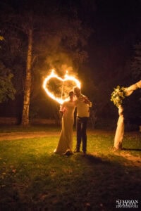 Романтичен нощен портрет на младоженци, прегърнати пред голямо пламтящо сърце по време на огнено шоу, заснет от сватбен фотограф Симеон Сълов.