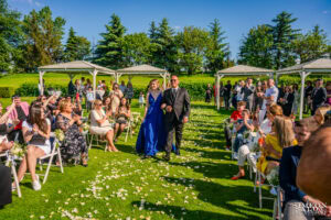 Младоженецът върви по пътеката към арката, воден от своята дъщеря в синя рокля, докато гостите ръкопляскат на сватба в Голф клуб "Св. София". Кадър от сватбен фотограф Симеон Сълов.