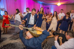 Музикант с бузуки свири на пода, докато младоженецът Радослав и гостите играят гръцки танц (сиртаки) около него на сватбата в хотел Маринела, кадър на професионален сватбен фотограф Симеон Сълов.