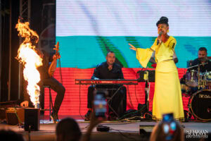 Петя Панева пее на сцената на Слатина Фест 2025, докато музикант до нея прави огнено шоу с горящ инструмент, заснета от професионален фотограф Симеон Сълов.