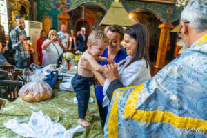 Майката и кръстницата предават разплакания Борис на свещеника преди потапянето в купела – емоционален момент, заснет от фотограф за кръщене.