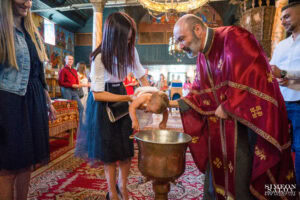 Свещеникът предава новокръстения Светозар в ръцете на неговата кръстница, която го поема с нова бяла кърпа след потапянето в купела.