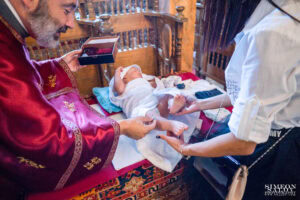 Свещеникът и кръстницата заедно обличат малкия Светозар след ритуала по Свето Кръщение, докато той лежи повит в бяла кърпа.