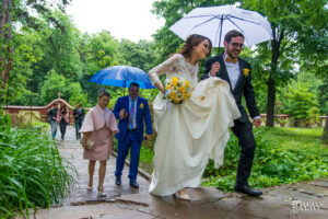 Щастливите младоженци Божин и Ханин вървят с чадър в дъжда в парка на Семинарията, а булката Ханин повдига роклята си. Кадър от сватбен фотограф Симеон Сълов.
