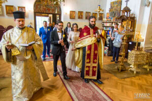 Младоженците Божин и Ханин, държащи запалени сватбени свещи, са водени от свещениците по време на църковния ритуал в храм "Свети Йоан Рилски". Кадър от **сватбен фотограф** Симеон Сълов.