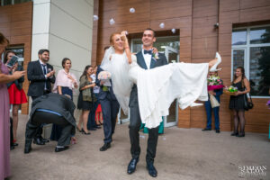 Младоженецът носи булката на ръце пред Обредна зала "Хиподрума", докато гостите ги посипват с конфети. Кадър от сватбен фотограф.