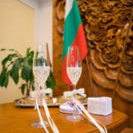 Ритуални чаши за шампанско и кутийка с халки на масата пред българския флаг в Обредна зала "Хиподрума", заснети от сватбен фотограф.