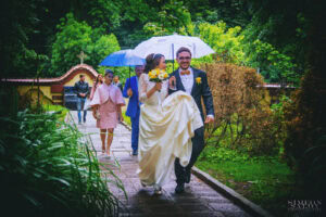 Щастливите младоженци Божин и Ханин вървят под чадъри в дъжда в парка на Семинарията. Романтичен кадър от сватбен фотограф София Симеон Сълов.