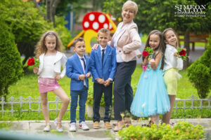 Детски фотограф за дипломиране: Група деца - момчета в костюми и момичета в рокли - позират със своята усмихната учителка в двора. Кадър от фотограф Симеон Сълов.