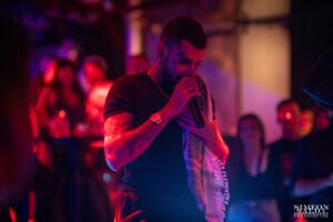 Емоционален и въздействащ кадър на Венци Венц (Venci Venc) по време на музикално събитие в София, заснет от Симеон Сълов. Изпълнителят е уловен в близък план с микрофон и кариран шал под контрастно червено и синьо клубно осветление.