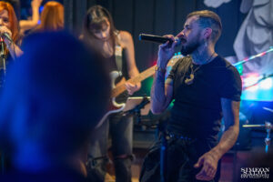 Професионална парти фотография на Венци Венц (Venci Venc), заснета от Симеон Сълов в нощен клуб в София. Изпълнителят пее на микрофон в близък план, докато на заден план се вижда китаристка с бяла електрическа китара под динамично синьо осветление.